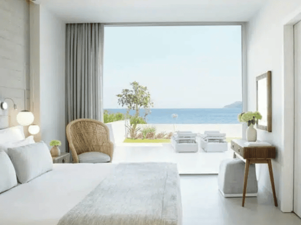 Sea view one-bedroom suite at Ikos Aria with private garden, featuring a bed, wicker chair, and large window overlooking the Aegean Sea.