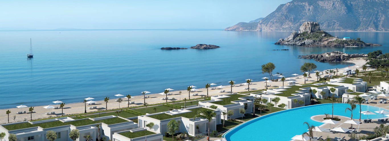 Ikos Aria resort on Kos, Greece, featuring modern white buildings, a beachfront on the Aegean Sea, a large pool, and a mountainous background.