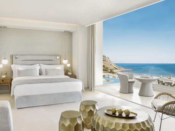 Interior of Ikos Aria Panorama Junior Suite with modern decor, a king-sized bed, and a balcony overlooking the sea and pool.
