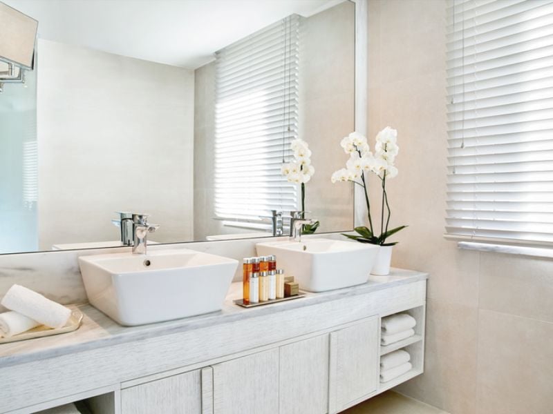Luxurious bathroom at Ikos Aria with dual sinks, white orchids, toiletries, and a large window with blinds.