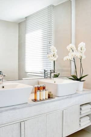 Luxurious bathroom at Ikos Aria with dual sinks, white orchids, toiletries, and a large window with blinds.