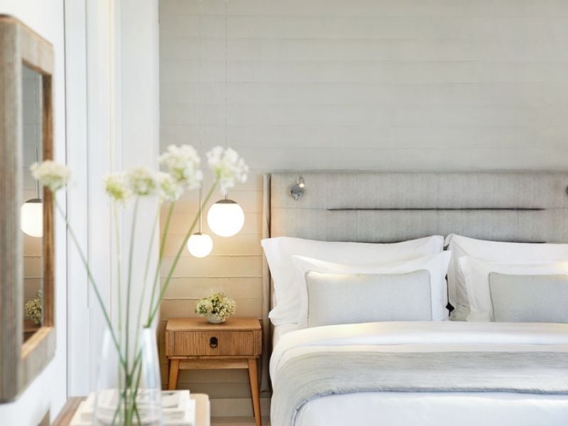 Elegant bedroom at Ikos Aria hotel with a double bed, light grey linens, a wooden nightstand, soft ambient lighting, and a mirror.