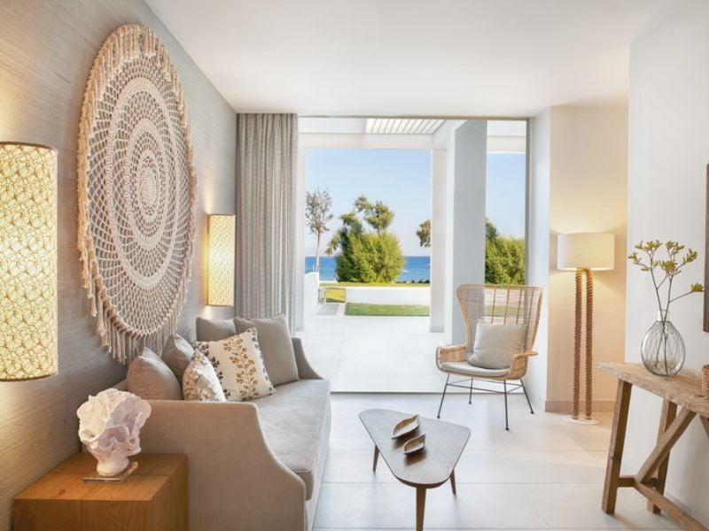 Ikos Aria living room with a sofa, decorative cushions, woven wall hanging, wooden table and chair, and large windows with sea views.