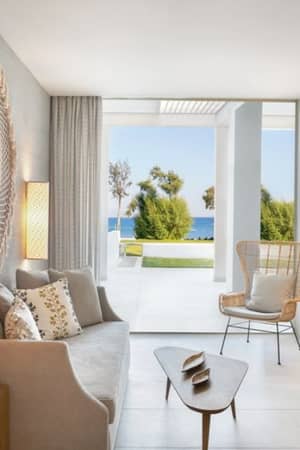 Ikos Aria living room with a sofa, decorative cushions, woven wall hanging, wooden table and chair, and large windows with sea views.