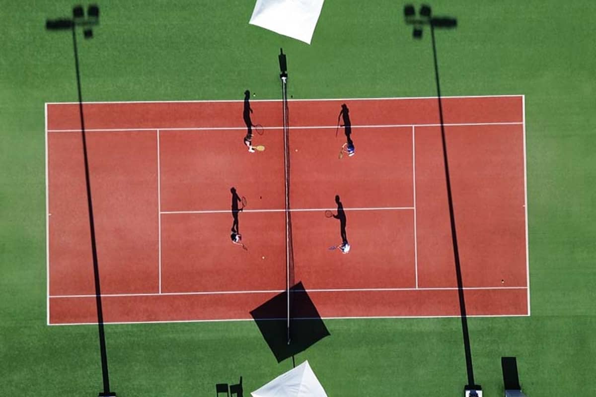 Aerial view of a tennis match on a red clay court at Ikos Aria, with players and shadows visible under white parasols.