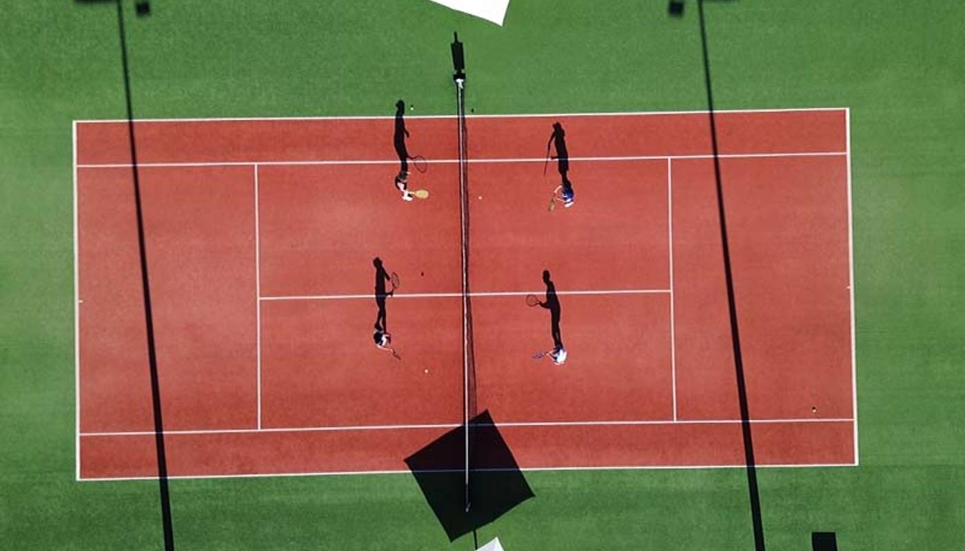 Aerial view of a tennis match on a red clay court at Ikos Aria, with players and shadows visible under white parasols.