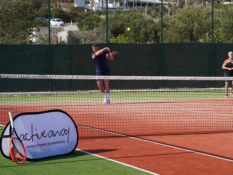 Tennis coach instructing a player on a clay court at Ikos Aria, with 'Active Away Unique Tennis Experiences' banner in the foreground.