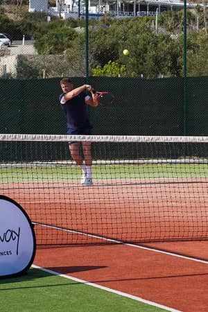 Tennis coach instructing a player on a clay court at Ikos Aria, with 'Active Away Unique Tennis Experiences' banner in the foreground.