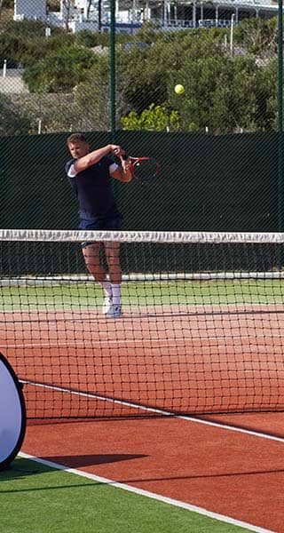 Tennis coach instructing a player on a clay court at Ikos Aria, with 'Active Away Unique Tennis Experiences' banner in the foreground.