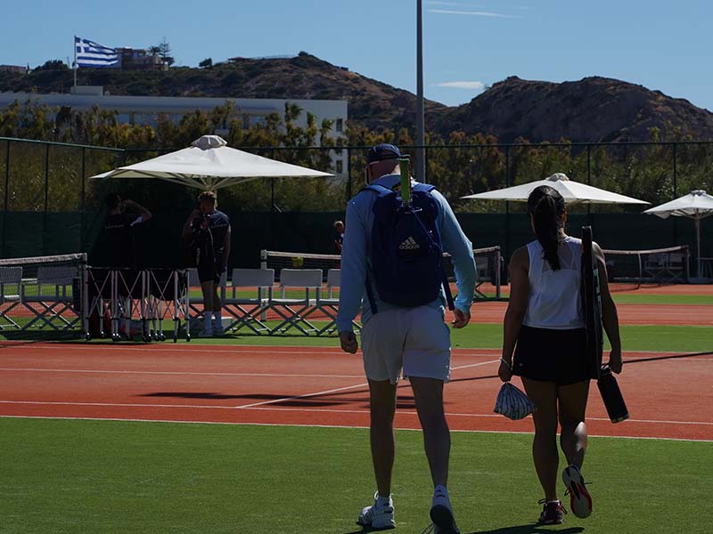 Tennis players walking towards a clay court at Ikos Aria resort with the Greek flag visible in the background.