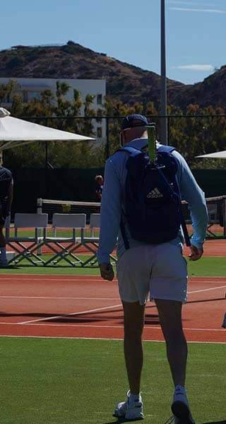Tennis players walking towards a clay court at Ikos Aria resort with the Greek flag visible in the background.