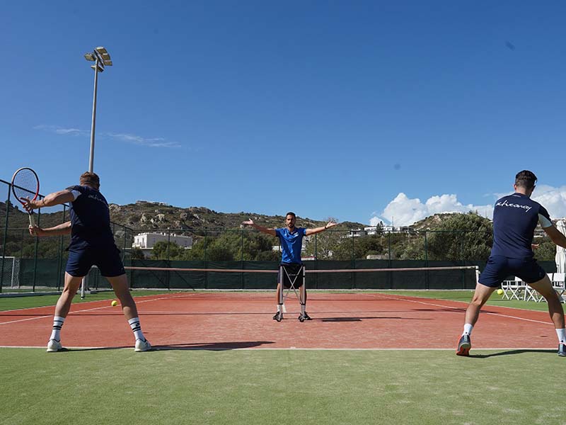 Outdoor tennis practice session with two players and a coach at the Ikos Aria resort, against a backdrop of hills.