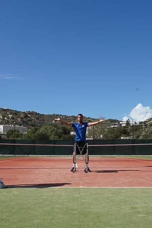 Outdoor tennis practice session with two players and a coach at the Ikos Aria resort, against a backdrop of hills.