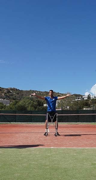 Outdoor tennis practice session with two players and a coach at the Ikos Aria resort, against a backdrop of hills.