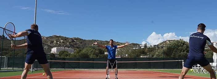 Outdoor tennis practice session with two players and a coach at the Ikos Aria resort, against a backdrop of hills.