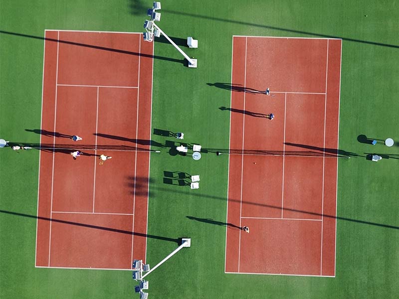 Aerial view of two clay tennis courts at Ikos Aria resort, with players and shadows on a green grass background.