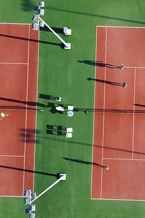 Aerial view of two clay tennis courts at Ikos Aria resort, with players and shadows on a green grass background.