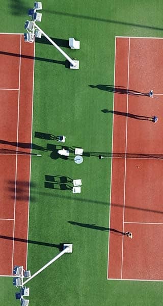 Aerial view of two clay tennis courts at Ikos Aria resort, with players and shadows on a green grass background.