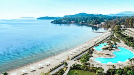Aerial view of Ikos Dassia luxury resort in Corfu, Greece, featuring a beach with parasols, an infinity pool, and lush gardens by the Ionian Sea.
