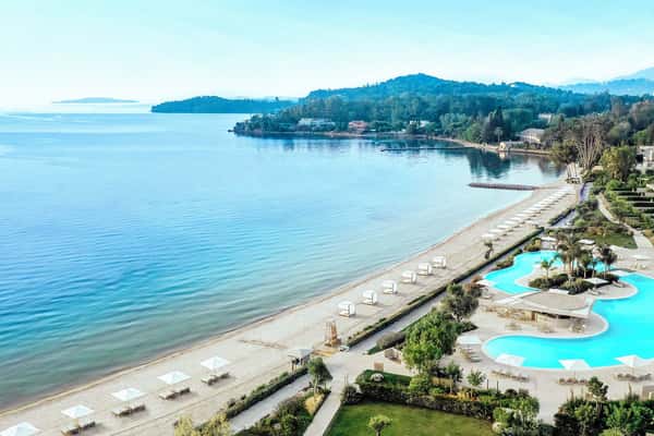 Aerial view of Ikos Dassia luxury resort in Corfu, Greece, featuring a beach with parasols, an infinity pool, and lush gardens by the Ionian Sea.