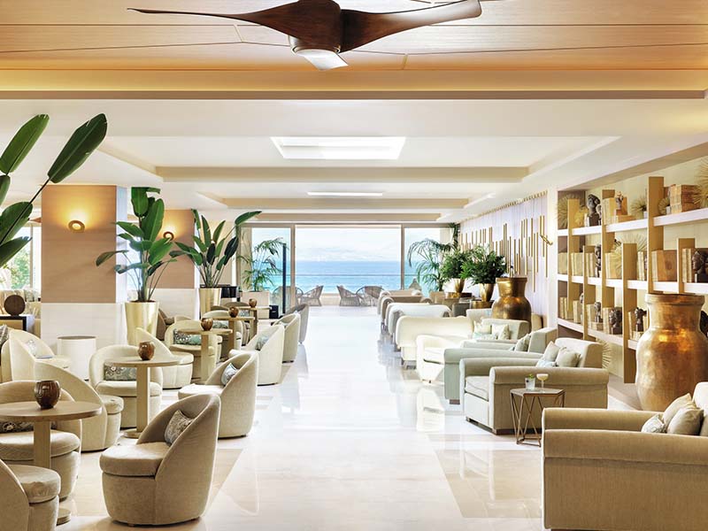 Hotel lounge at Ikos Dassia with beige sofas, large windows showcasing an ocean view, and elegant decor including plants and vases.
