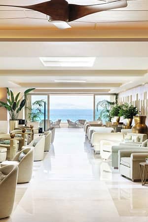 Hotel lounge at Ikos Dassia with beige sofas, large windows showcasing an ocean view, and elegant decor including plants and vases.