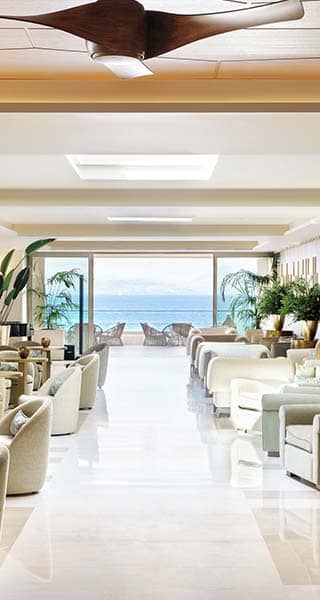 Hotel lounge at Ikos Dassia with beige sofas, large windows showcasing an ocean view, and elegant decor including plants and vases.