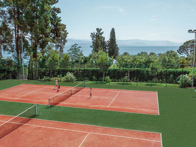 Outdoor tennis courts at Ikos Dassia, bordered by greenery with a sea view, featuring players in a match under a clear blue sky.