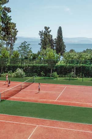 Outdoor tennis courts at Ikos Dassia, bordered by greenery with a sea view, featuring players in a match under a clear blue sky.