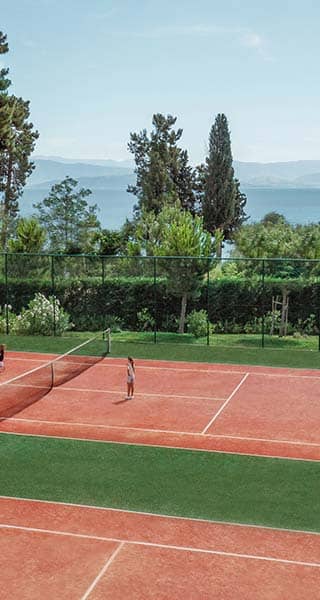 Outdoor tennis courts at Ikos Dassia, bordered by greenery with a sea view, featuring players in a match under a clear blue sky.