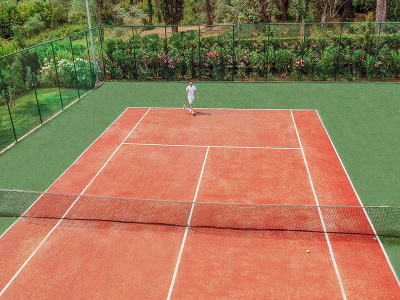 Tennis player on a red clay court at Ikos Dassia, surrounded by greenery.