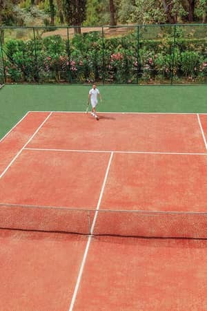Tennis player on a red clay court at Ikos Dassia, surrounded by greenery.