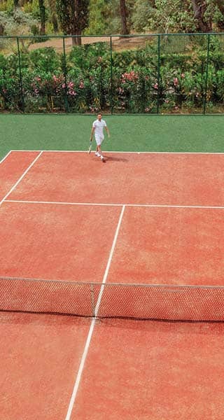 Tennis player on a red clay court at Ikos Dassia, surrounded by greenery.