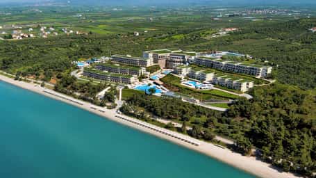 Aerial view of Ikos Oceania resort in Greece, with modern buildings, pools, beachfront, and green surroundings.