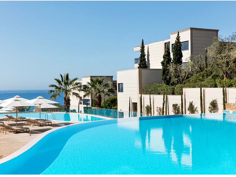 View of Ikos Oceania resort's infinity pool surrounded by sun loungers, parasols, modern buildings, lush greenery, and the sea in the background.