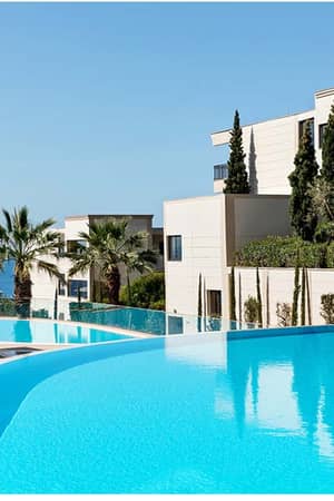 View of Ikos Oceania resort's infinity pool surrounded by sun loungers, parasols, modern buildings, lush greenery, and the sea in the background.