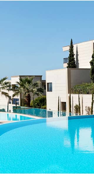 View of Ikos Oceania resort's infinity pool surrounded by sun loungers, parasols, modern buildings, lush greenery, and the sea in the background.