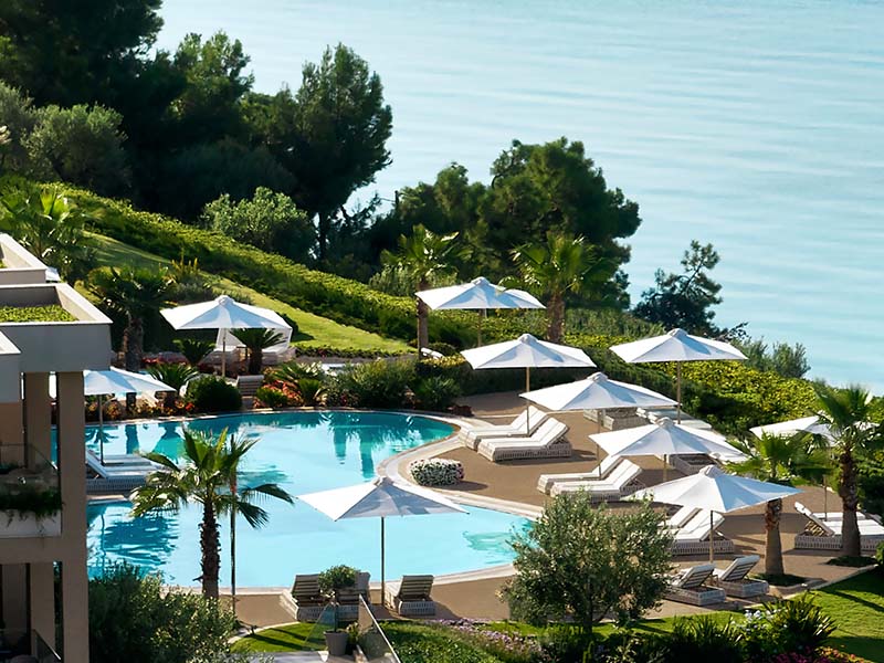 Aerial view of Ikos Oceania resort's poolside area with sun loungers, white parasols, lush greenery, and the Mediterranean Sea.