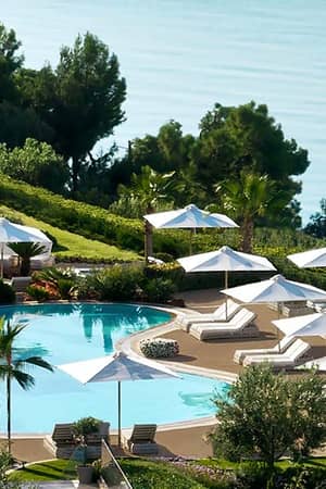 Aerial view of Ikos Oceania resort's poolside area with sun loungers, white parasols, lush greenery, and the Mediterranean Sea.
