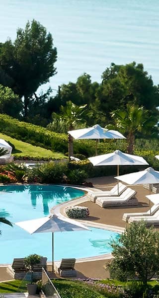 Aerial view of Ikos Oceania resort's poolside area with sun loungers, white parasols, lush greenery, and the Mediterranean Sea.