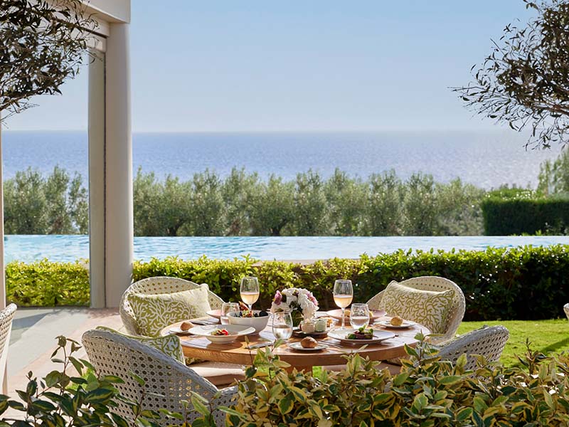 Outdoor dining area at Ikos Oceania with a table set for four, offering views of an infinity pool and the Aegean Sea.