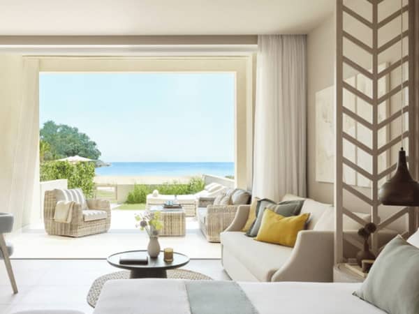 Deluxe Junior Bungalow Suite at Ikos Odisia, featuring a private garden and sea view through large sliding doors, with elegant interior seating and decor in soft colours.