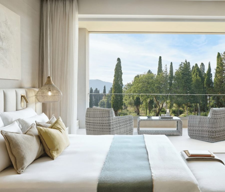 Interior of Ikos Odisia Deluxe One Bedroom Bungalow Suite, showing a bed with pillows and a balcony offering a garden view with wicker chairs.