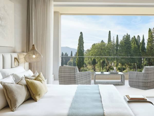 Interior of Ikos Odisia Deluxe One Bedroom Bungalow Suite, showing a bed with pillows and a balcony offering a garden view with wicker chairs.