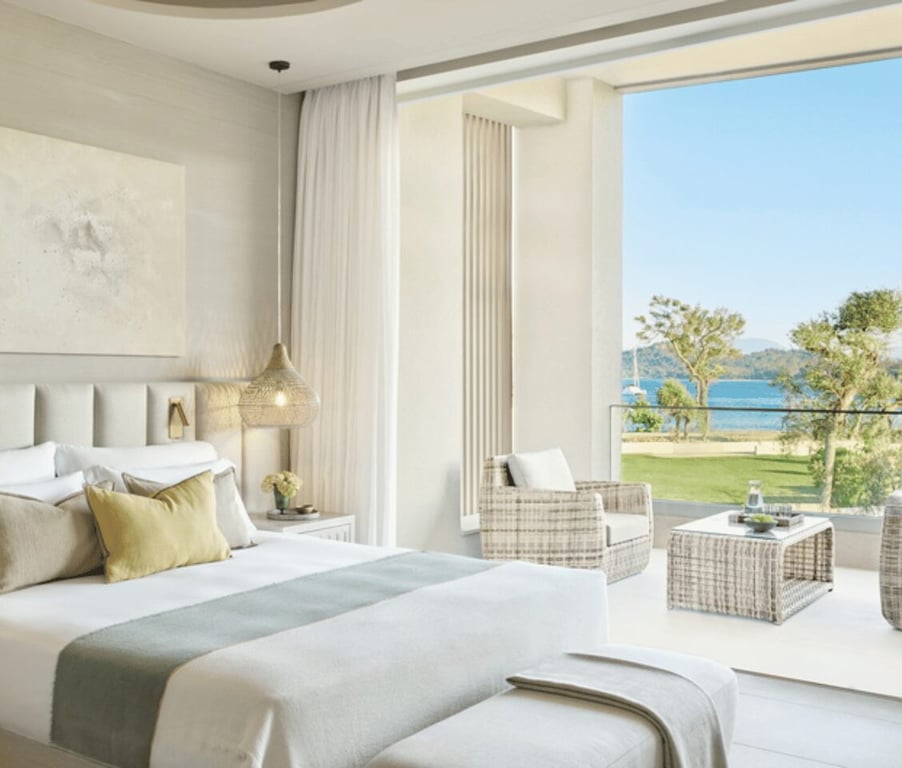 Interior of Ikos Odisia deluxe one-bedroom bungalow suite with balcony and sea view, featuring a stylish bed and wicker balcony furniture.