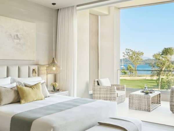Interior of Ikos Odisia deluxe one-bedroom bungalow suite with balcony and sea view, featuring a stylish bed and wicker balcony furniture.