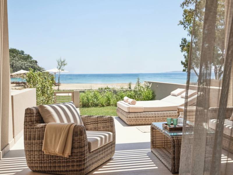 View from Ikos Odisia balcony showing wicker furniture, a lush garden, and a scenic beach with a blue sea backdrop.