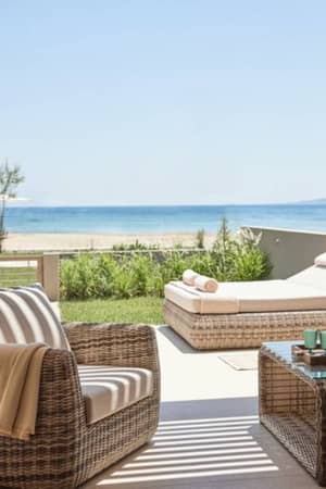 View from Ikos Odisia balcony showing wicker furniture, a lush garden, and a scenic beach with a blue sea backdrop.