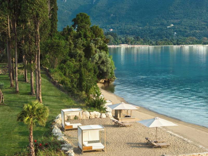 Ikos Odisia Beach with sun loungers and umbrellas, lush greenery, and mountains in the background.