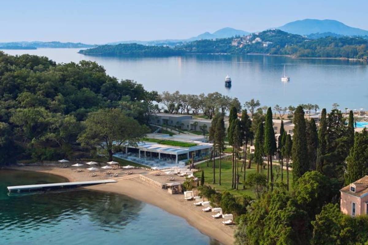 Aerial view of Ikos Odisia resort with sandy beach, sun loungers, green gardens, and a beachfront pavilion nestled in a coastal bay.